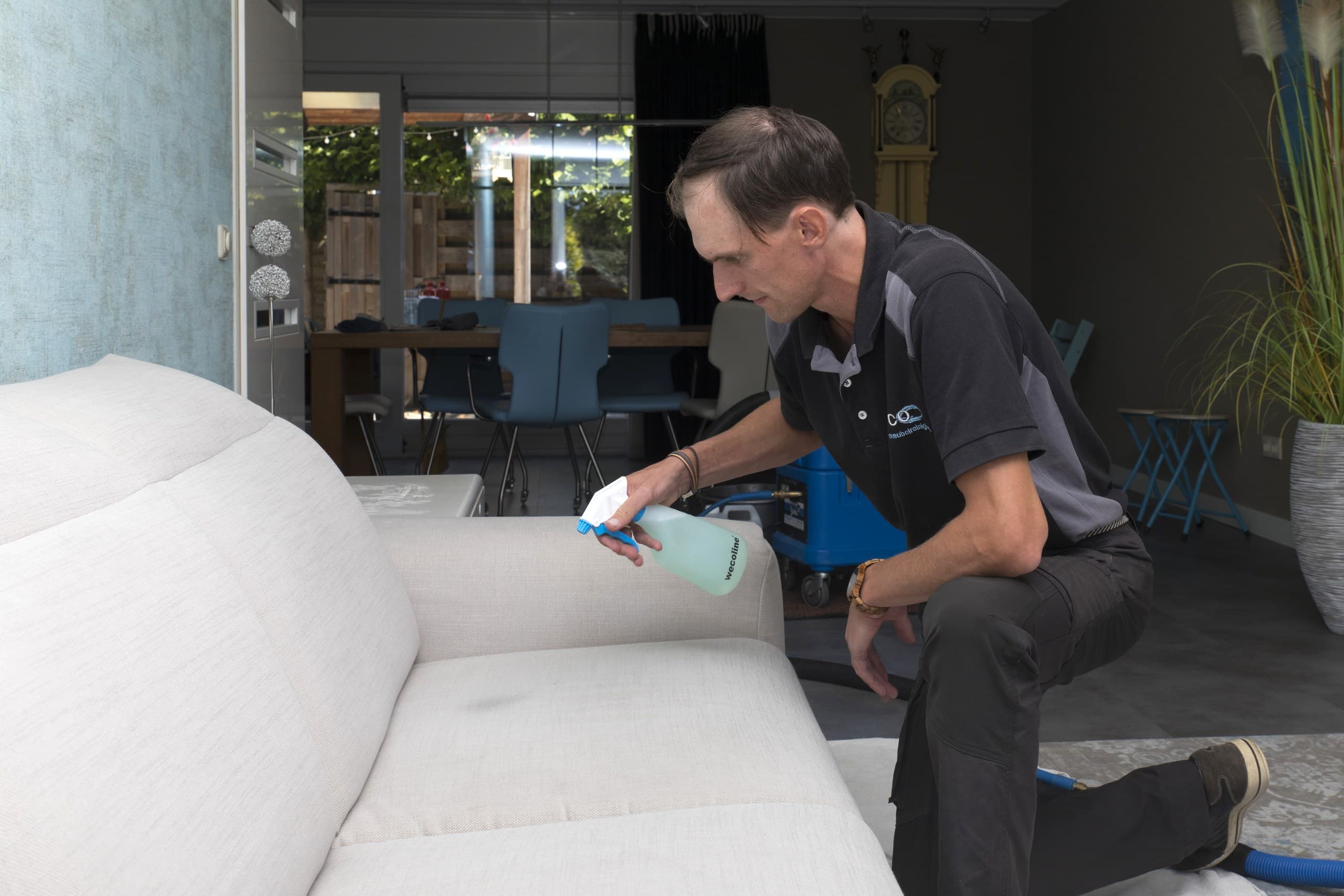 Medewerker brengt reinigingsmiddel aan op een lichte stoffen bank in een woonkamer