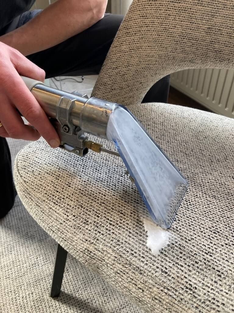 Hand reinigt beige stoelstof met sproei-extractiemondstuk, schuim zichtbaar op zitting